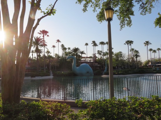 Apatosaurus statue in a theme park lagoon, attached to a snack shop. Sun rises on the left; water reflection is pretty.