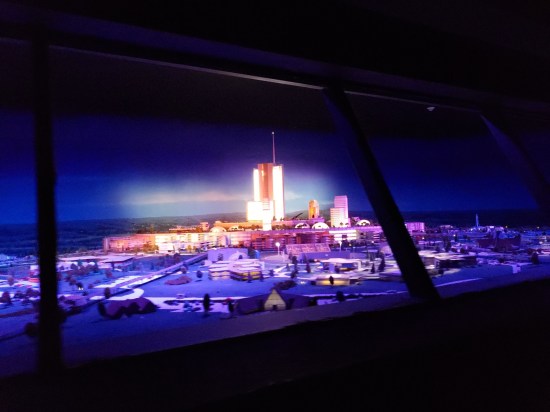 Diorama of what looks like a futuristic shopping mall. All around the diorama is darkness.