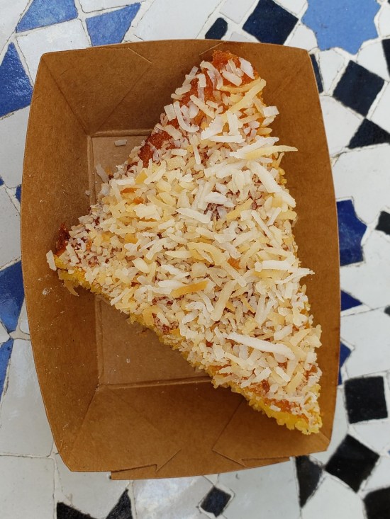 Triangular cake slice topped with finely grated cheeses and fruit filling, served in a small rectangular cardboard restaurant bowl.