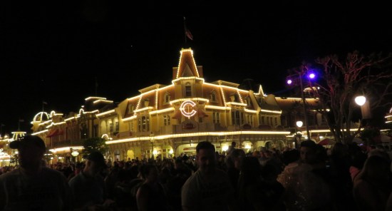 Old shops lit up at night, one with a giant C on the front. Shadowy people pack the streets.