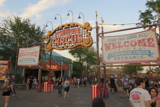 Sign over Disney World walkway says "Storybook Circus". Welcome signs flank either side. Park lights are on but the sky is still daylight blue.