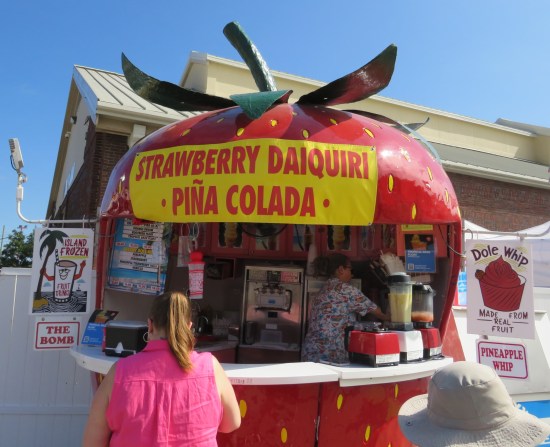 Drink stand shaped like a giant strawberry with one lady making a drink inside. Signs offer strawberry daiquiris, piña coladas, Dole whips, and some fruit drink called "The Bomb".