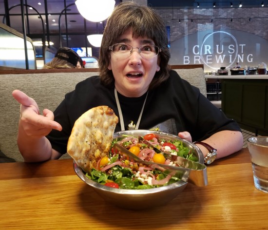 Anne sitting in a gastropub booth point at her lunch, a salad served in a giant metal mixing bowl.