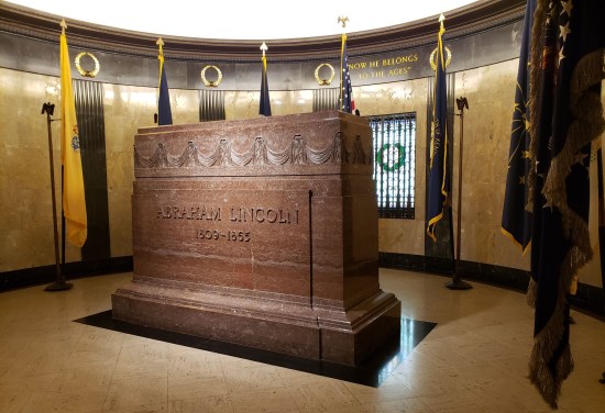 Indoor brown rectangular monument shaped like a tall sarcophagus. Inscribed on the front: "Abraham Lincoln 1809-1865". State flags line the curved yellow wall behind it, plus the quote "Now he belongs to the ages."