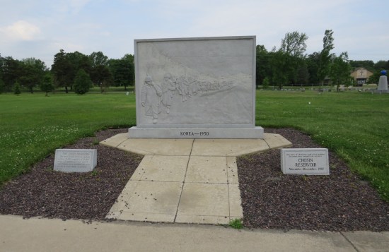 Large concrete standalone wall etched with soldiers walking in a line, labeled "Korea 1950". The wall stands at the end of a short sidewalk and is surrounded by a rectangular gravel bed.