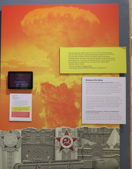 Museum narration placards in front of a wall-size photo of a nuclear explosion, recolored orange.