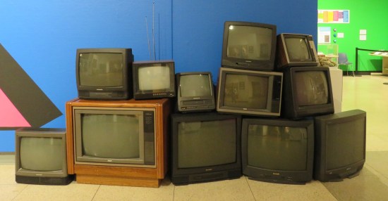 A dozen CRT TVs in various sizes stacked against a blue wall.