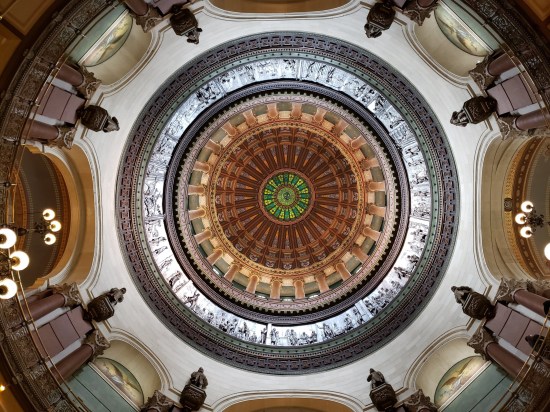Looking up inside a capitol dome. Brown and tan stained glass middle ring, green center circle, circular silvery frieze in the outer circle. Around the perimeter are archways to various halls.