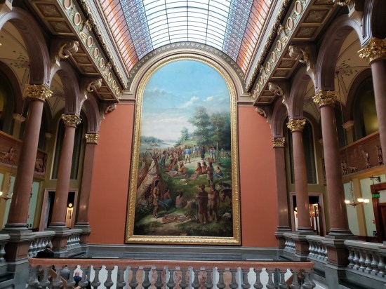 Large painting with arched top. One white guy speaks in the middle of a large Native tribe. Other white folks in his party hang back. Sunny outdoors, river and woods in the distance.