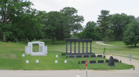 Two giant family plots side by side. One is a black Greek column with two long rows of headstones, mostly empty for now. The other is a large white open double-doorway with its fake doors opened. Six white headstones are spaced apart in front.