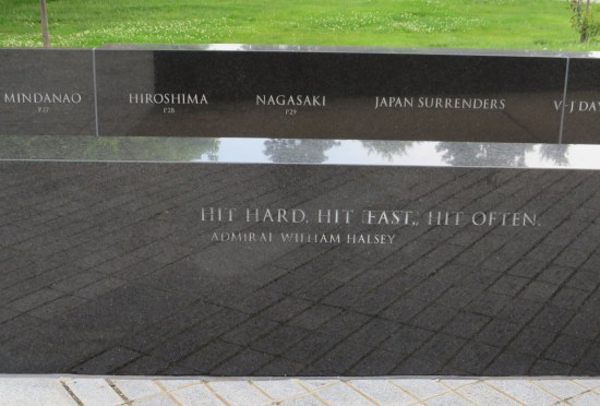 Short black marble wall engraved with quote: "Hit hard. Hit fast. Hit often." Behind it, a second wall is engraved with a timeline of WWII events -- Hiroshima, Nagasaki, and Japan's surrender.