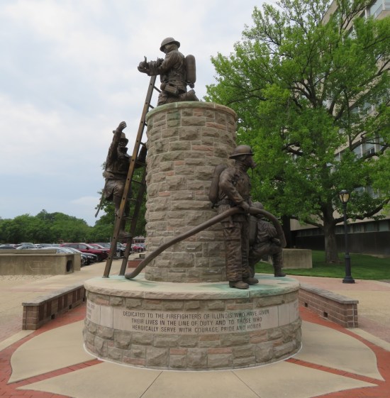 Sculpture of two firemen climbing a ladder up a factory smokestack while two other firemen wrangle a fire hose around the base.