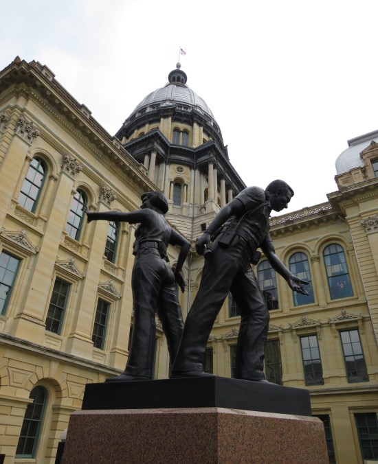 Statue of one male police officer lending a hand and one female officer stopping traffic, standing behind the Illinois State Capitol.