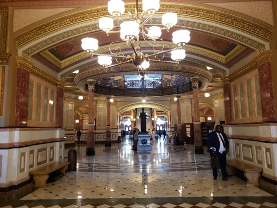 Interior architecture, lots of tan and brown stonework leading to the rotunda. Chandeliers, white wainscotting, white marble floor with gray geometric patterns. In the distance is a statue and a staircase.