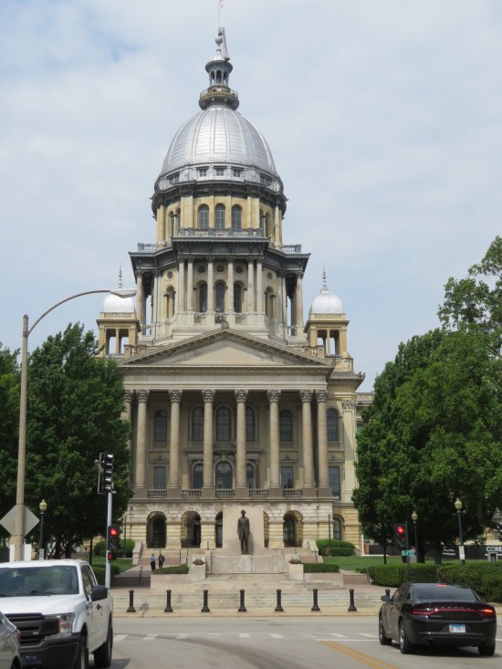 Domed state capitol building with trees on either side and a thin Lincoln statue in front.