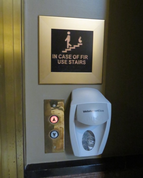 Misspelled sign "In Case of Fir" with a tiny staircase symbol. Hung above elevator buttons and a hand sanitizer dispenser.