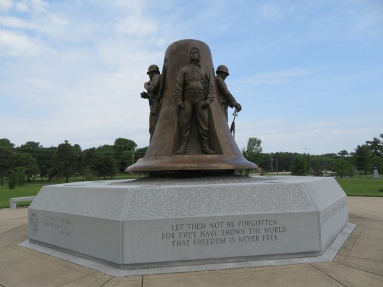 Large metal bell with four soldiers standing on it, mounted on a stone pedestal. Message engraved on one side: "Let them not be forgotten, for they have shown the world that freedom is never free."