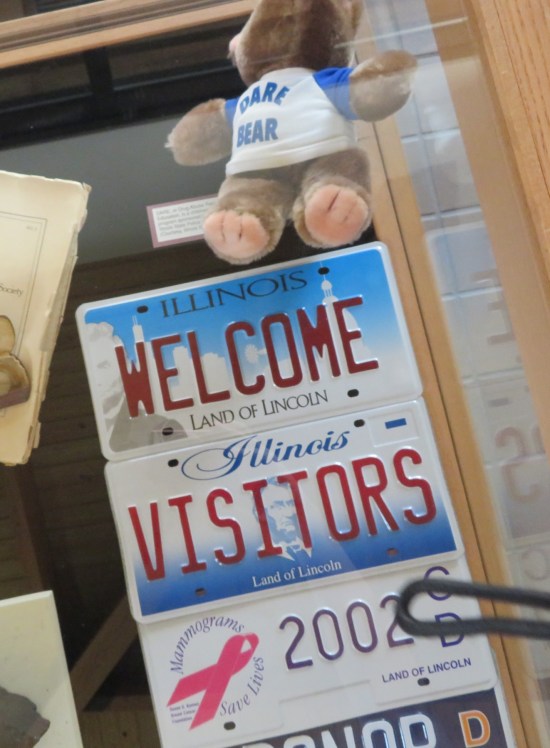 Vitrine containing Illinois license plates reading "Welcome" and "Visitors". Also, a teddy bear with a shirt reading "DARE BEAR".