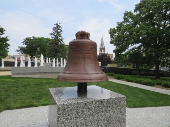 Small bronze Liberty Bell replica on a pedestal. Far behind are rows of fountains.