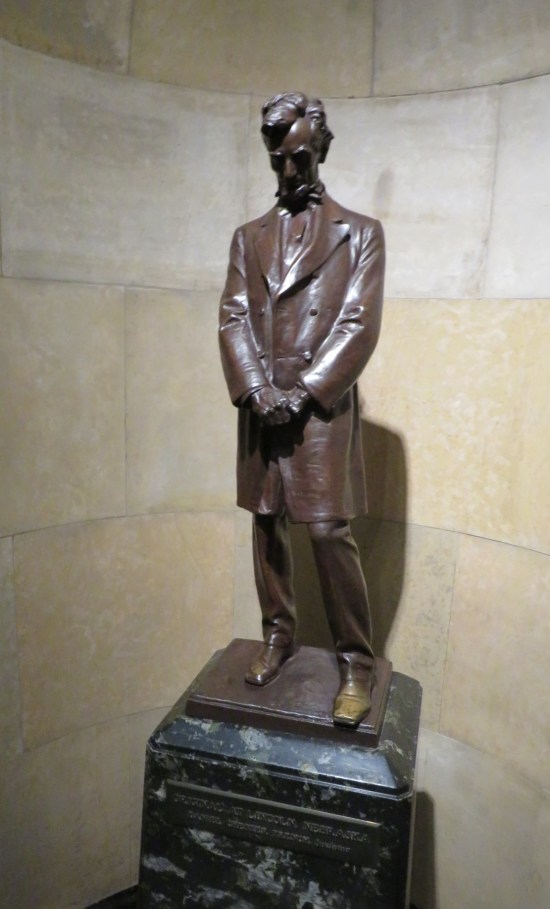 Statue of Lincoln with head bowed and hands clasped at his waist, wearing a long dress jacket.