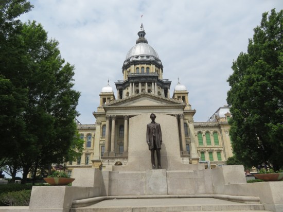 Thin Lincoln statue on a staircase landing in front of a capitol building. Lincoln is young, beardless, and wearing a long coat with his arms down at his sides.