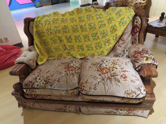 Old brown love seat with comfy cushions, sparse wildflower pattern, and an ugly yellow quilt thrown over it.