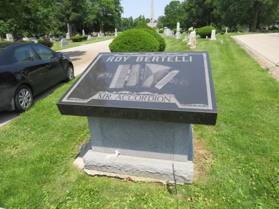 Black crypt marker for Roy Bertelli, Mr. Accordion, with a picture of an accordion, sitting on a cemetery roadway median. My car is parked by the curb.