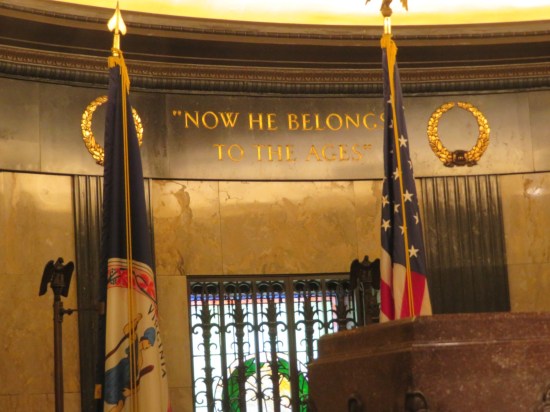 Etched in the wall behind Lincoln's monument are the words "Now he belongs to the ages." Flags flank each side. Lighting is yellowish.