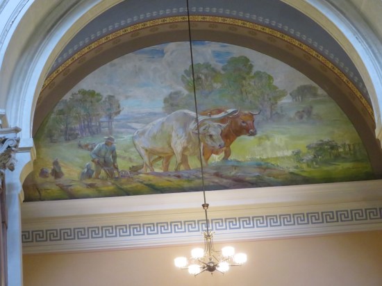 Half-circle painting of a farmer with oxen plowing uphill while two women sow seeds behind them.