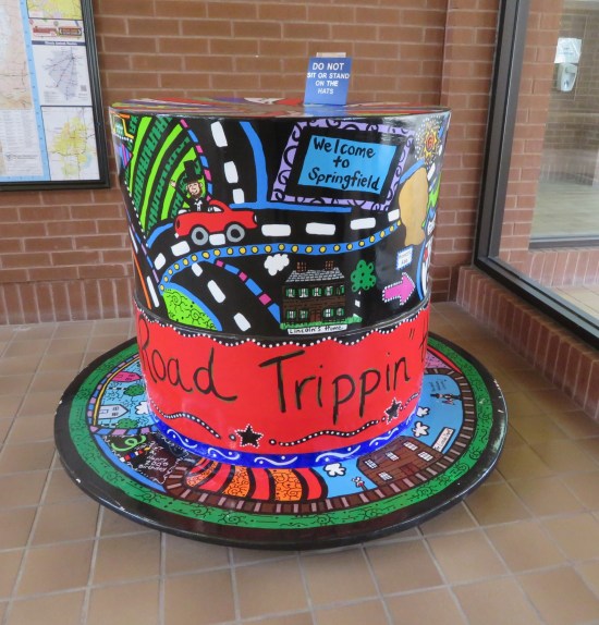 Colorful 5-foot-tall top hat in a visitors' center. Images on the hat include young Abe Lincoln riding in a red car on a highway, a "Welcome to Springfield" sign, the official Lincoln's Home museum, and a wraparound cursive logo starting with the words "Road Trippin'".