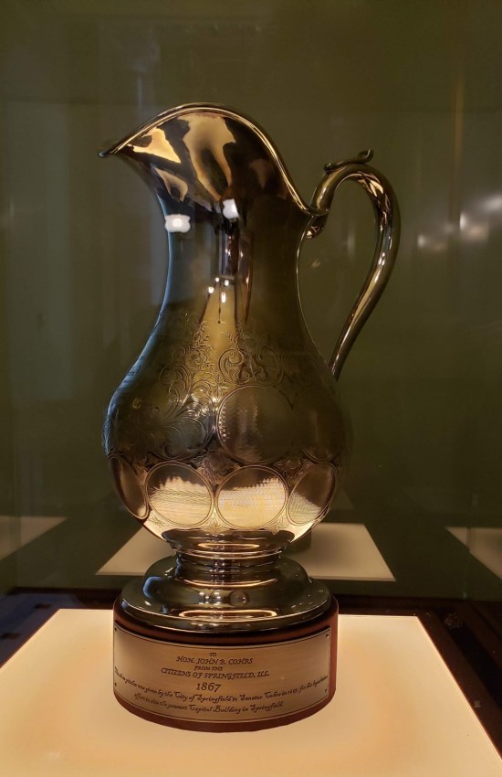Silver pitcher in a vitrine. The caption summarizes its inscription. My reflection is distorted.