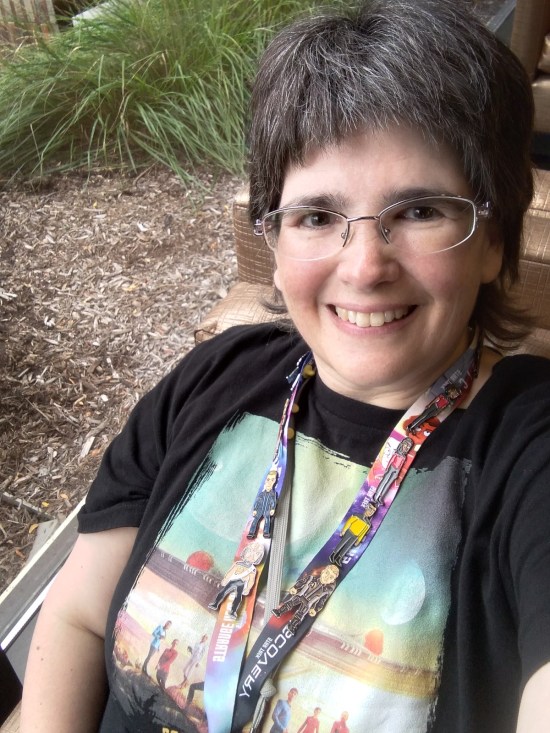An Anne selfie with her "Strange New Worlds" T-shirt and lanyard covered with Trek character pins.
