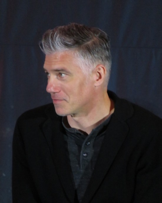Anson Mount on stage, dressed darkly, listening patiently.
