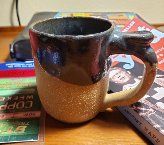 Brown and beige ceramic mug, sitting next to my con badge and Anthony Rapp's book.