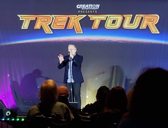 Anthony Rapp signing on a stage. Blue backdrop has a large TREK TOUR logo with purple and white light arcs drawn beneath it.