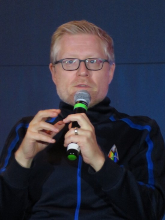 Anthony Rapp on stage with a mic, glasses, and a Pride-rainbow Trek Delta pin.