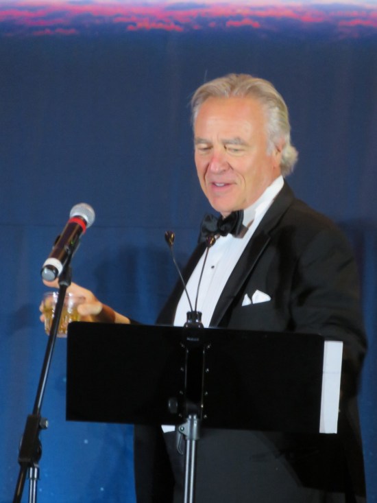 Casey Biggs in tuxedo with drink in hand onstage.