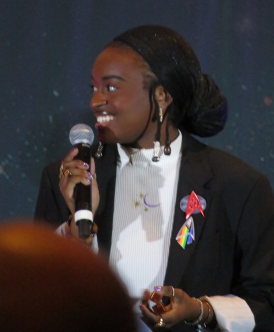 Celia Rose Gooding on stage wearing a jacket and two Trek Delta pins, including a Pride-themed one.