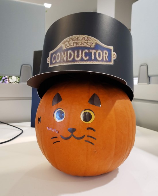Pumpkin with a cat face drawn on it. On top is a cardboard hat labeled "POLAR EXPRESS CONDUCTOR".