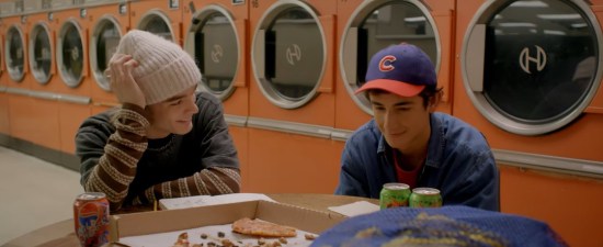 Two young guys at a laundromat table with a nearly finished pizza. Orange dryers line the walls.