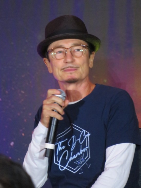 Dominic Keating holding a mic and wearing a bowler and a D-Con Chamber shirt.