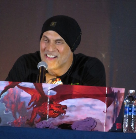 Todd Stashwick in black cap behind a Dungeon Master's screen with a red dragon painted on it.