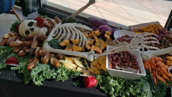 Halloween skeleton lying on a table surrounded by party snacks -- breadsticks, sausages, cheeses, carrots, and so on.