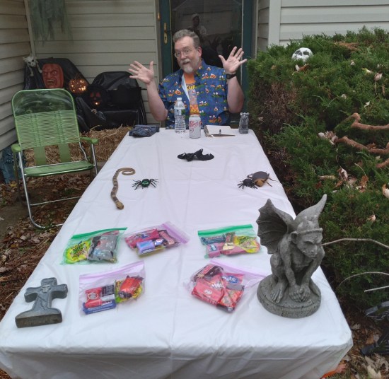 Me sitting and doing jazz hands at a yard sale table on our front sidewalk. On the table are Ziploc bags of candy and small Halloween critters and tiny tombstones for decoration.
