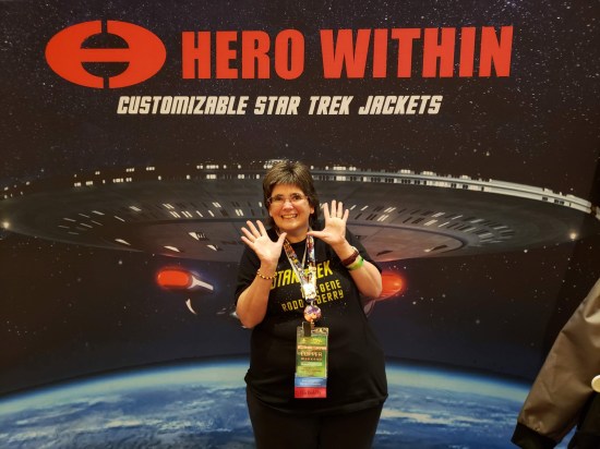 Anne doing jazz hands in front of a large Enterprise poster at the Hero Within Customizable Jackets booth.