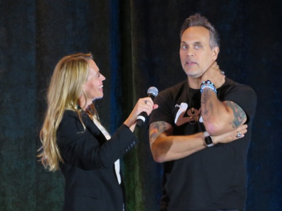 Jeri Ryan and Todd Stashwick sharing a stage. She holds the mic toward him as ne natters on.