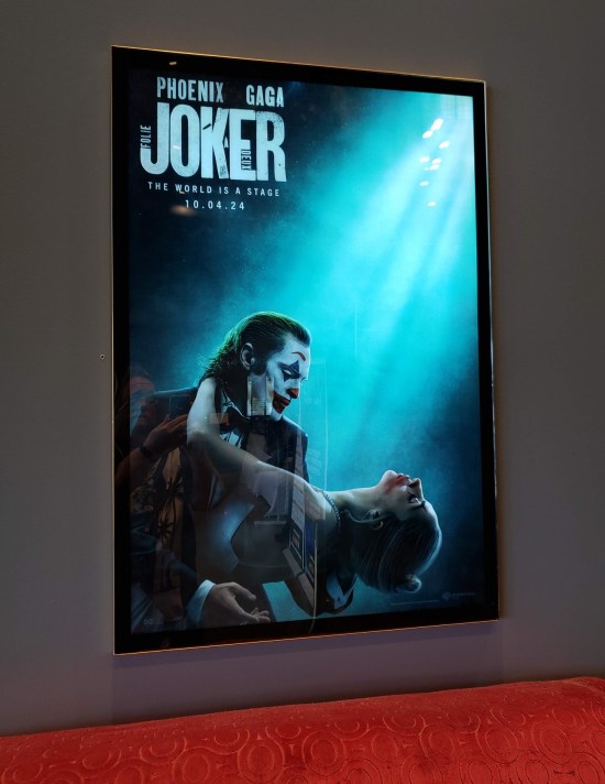 Poster for "Joker Folie a Deux" hanging in a theater lobby with gray walls above a red couch.