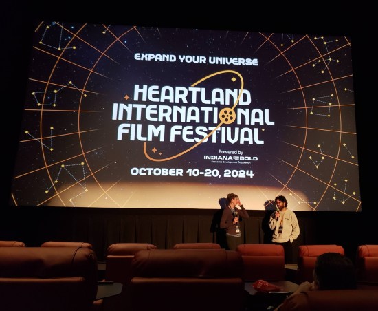 Heartland International Film Festival 2024 logo on a full movie screen with two people standing below it.