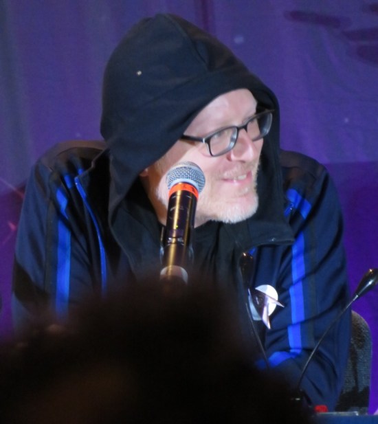Anthony Rapp in glasses and blue hoodie, with hood up.