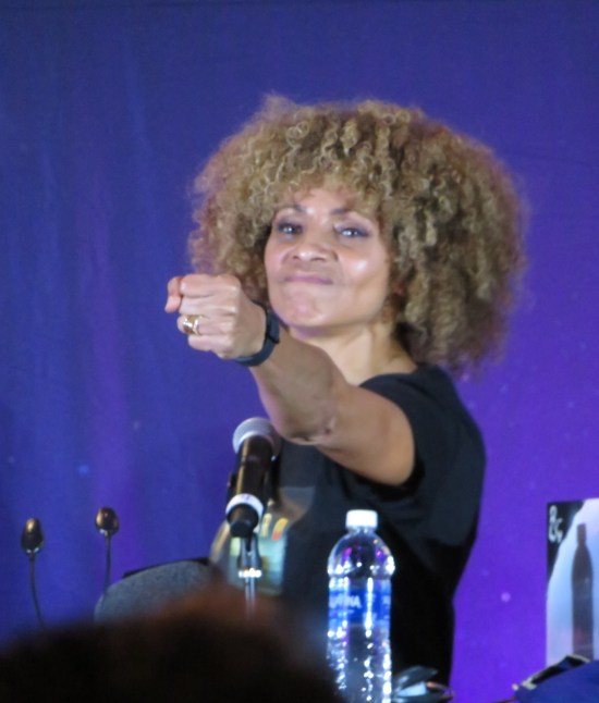 Michelle Hurd at a table in a Picard T-shirt, one arm extended forward as if holding an imaginary bow and arrow.